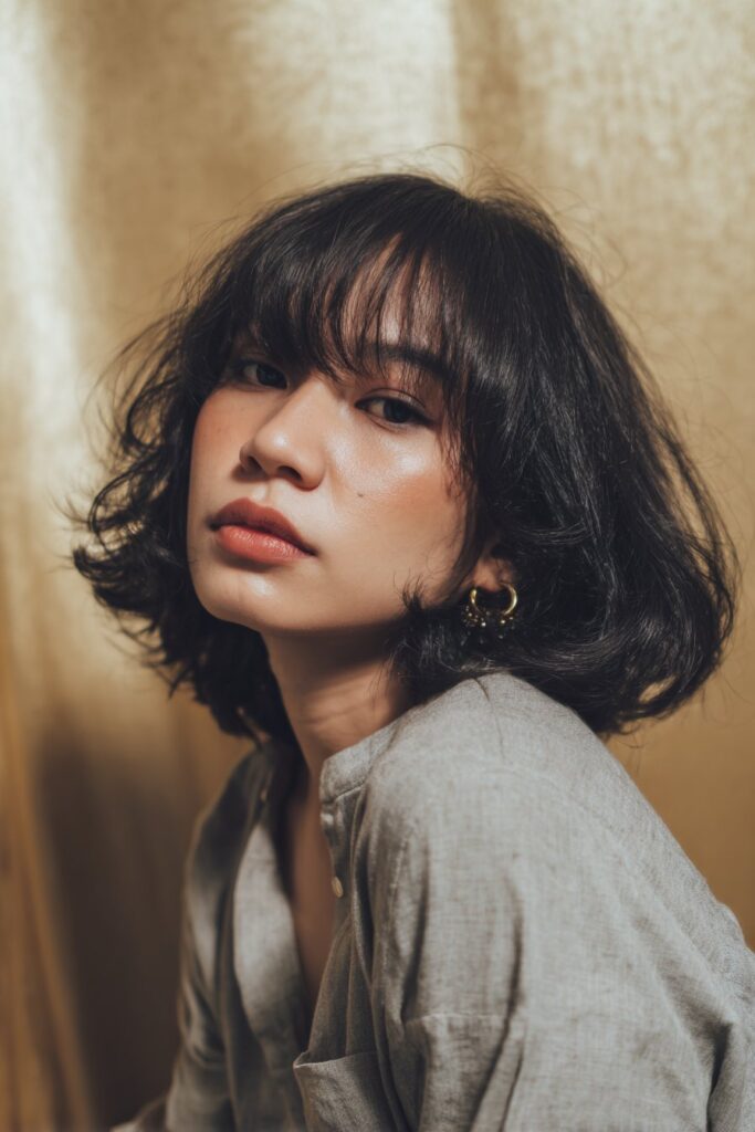 A stunning young woman in her 20s with natural skin tone, showcasing a bob hairstyle with side bangs featuring a feathered bob with side bangs, featuring a feathered bob with side-swept bangs, captured in a close-up shot focusing on the hair, photographed against a seamless light gold backdrop with a glossy sheen. She has subtle makeup, a chic outfit, realistic hair texture, warm skin tone, and a confident expression, captured with soft natural lighting and an Instagram-style beauty vibe.