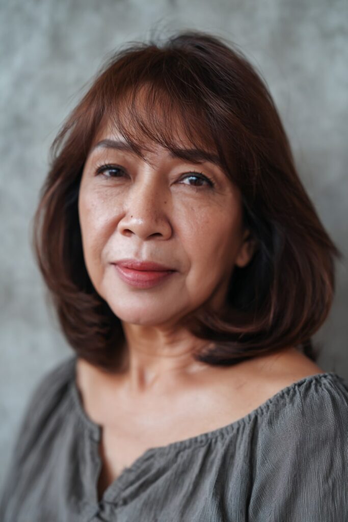 Woman over 50 with shoulder-length feathered shag, bangs, dark chestnut hair, gray backdrop.
