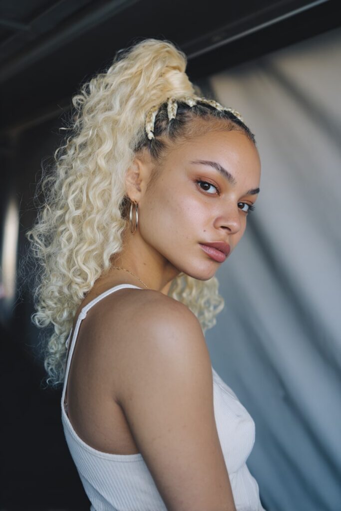 Black girl with half-up Barbie ponytail, quick weave curls, platinum blonde, gray backdrop.