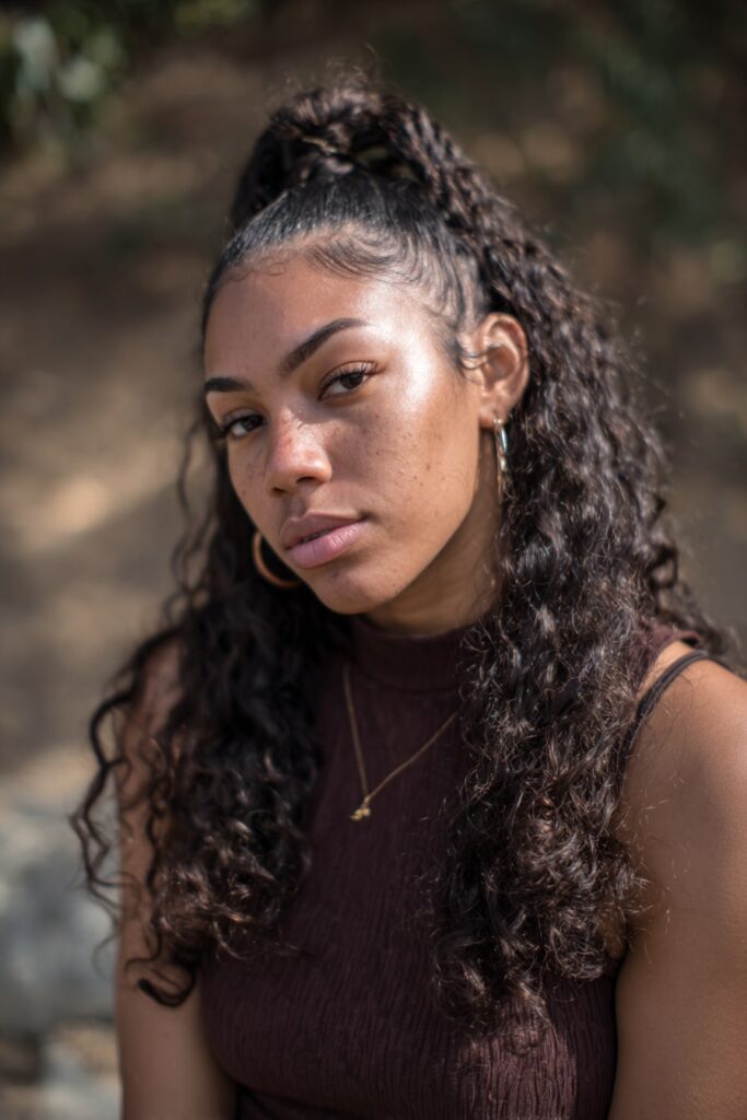 Black girl with half-up top knot, loose curls, clip-ins, dark brown, forest backdrop.