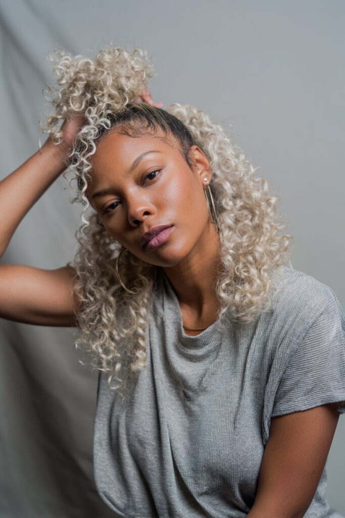Black girl with half-up 613 blonde wig, curly hairstyle, platinum blonde, gray backdrop.