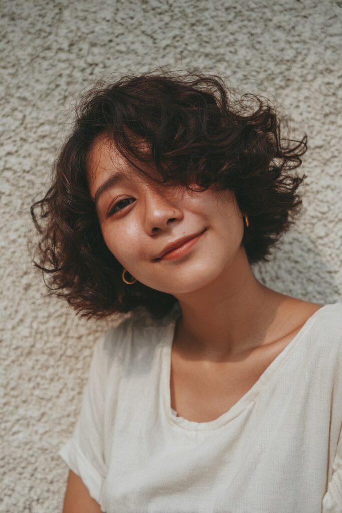 Woman with short half-updo, crimped texture, dark chestnut hair, plaster backdrop.