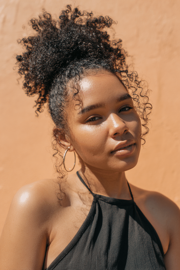 A stunning young woman in her 20s with natural skin tone, showcasing a homecoming hairstyle for curly hair with a high puff glam, featuring a high, voluminous puff with curly texture, captured in a close-up shot focusing on the hair, photographed against a seamless warm peach backdrop with a matte finish. She has subtle makeup, a chic homecoming dress, realistic hair texture, warm skin tone, and a confident expression, captured with soft natural lighting and an Instagram-style beauty vibe.