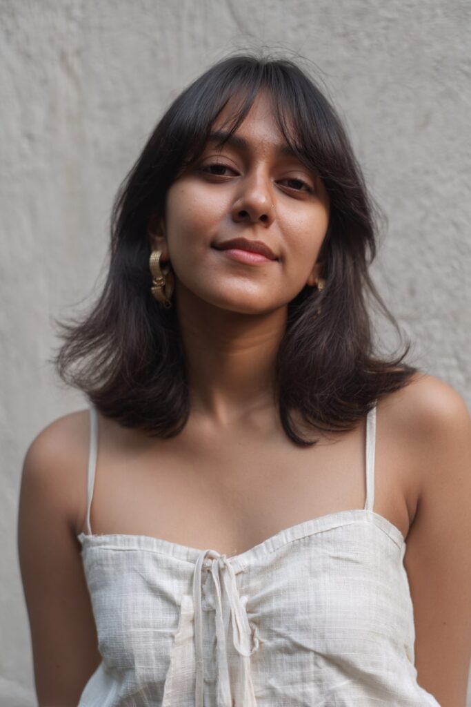 Young woman with medium-length layered waves, bottleneck bangs, natural black hair, textured plaster backdrop.