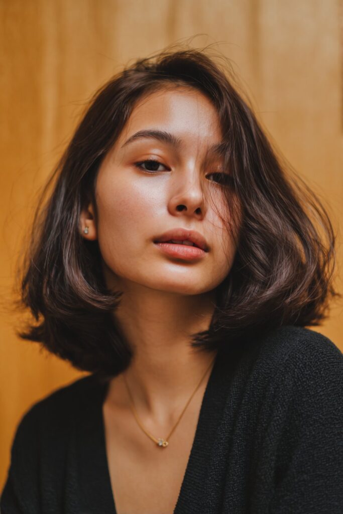 A stunning young woman in her 20s with natural skin tone, showcasing a bob hairstyle for going out featuring a middle part bob, featuring a bob with a clean middle part, captured in a close-up shot focusing on the hair, photographed against a seamless rich aubergine backdrop with a glossy texture for optimal color contrast. She has subtle makeup, a chic evening outfit, realistic hair texture, warm skin tone, and a confident expression, captured with soft natural lighting and an Instagram-style beauty vibe.