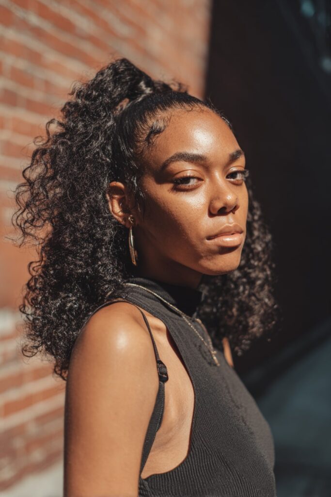 Black girl with half-up natural curls, clip-ins, natural black hair, brick backdrop.