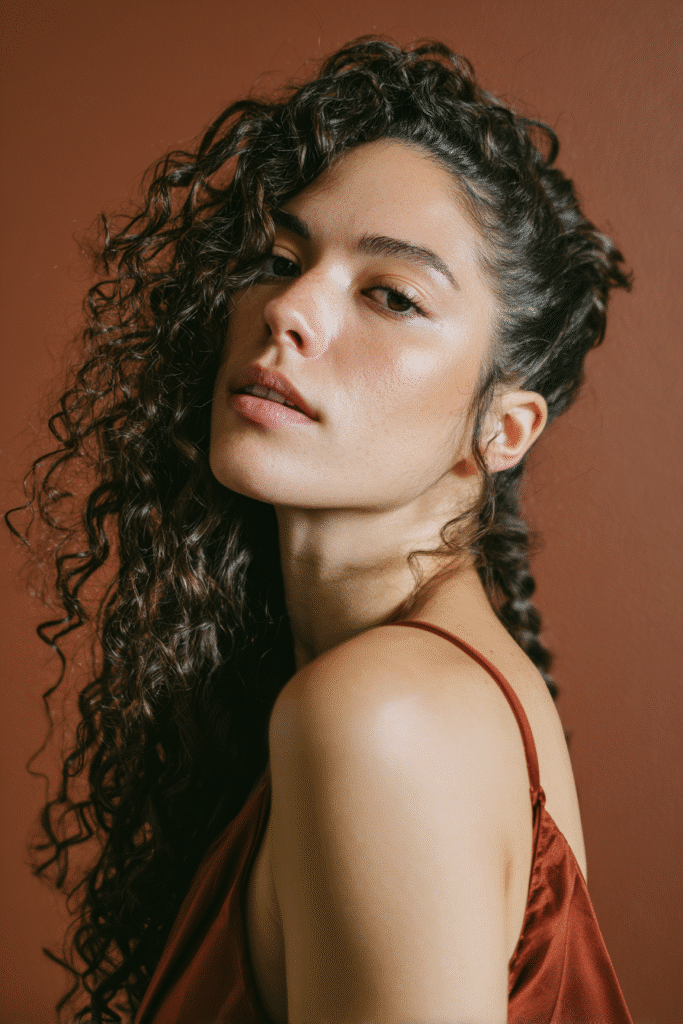 A stunning young woman in her 20s with natural skin tone, showcasing a homecoming hairstyle for curly hair with a romantic side braid, featuring a loose side braid with romantic curly waves, captured in a close-up shot focusing on the hair, photographed against a seamless warm terracotta backdrop with a satin finish. She has subtle makeup, a chic homecoming dress, realistic hair texture, warm skin tone, and a confident expression, captured with soft natural lighting and an Instagram-style beauty vibe.