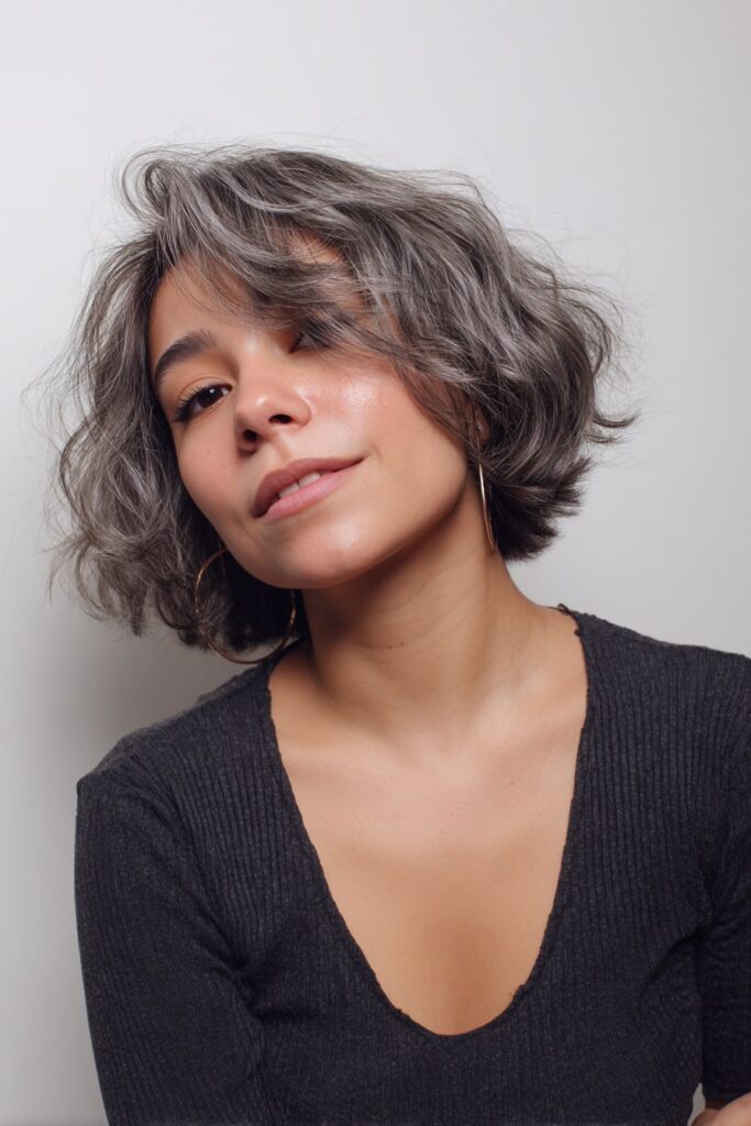 Young woman with medium-short tousled bob, side fringe, layers, natural gray hair, white backdrop.