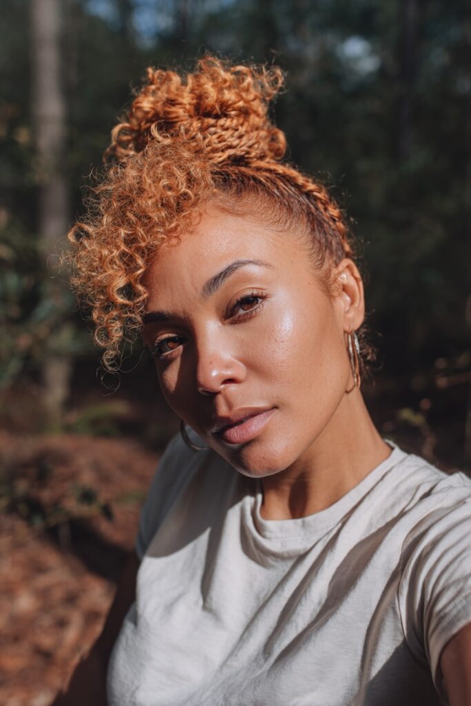 Woman with half up half down curly hair, tousled curls, braided halo, forest backdrop.