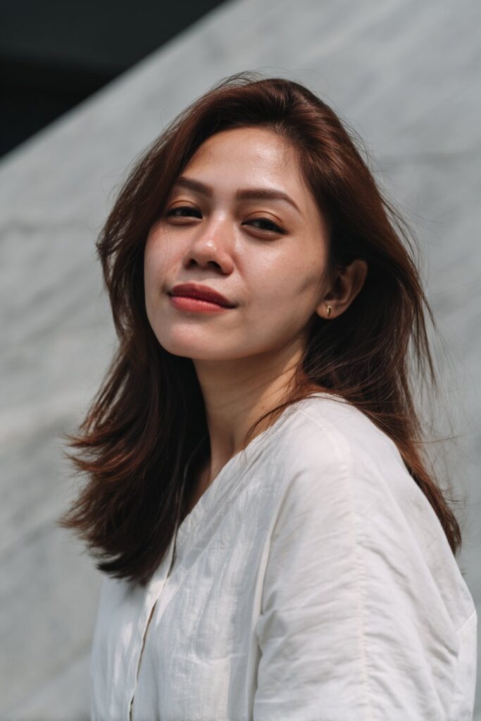 A stylish American woman in her 20s with a sleek angled lob haircut, featuring longer front layers and subtle volume, photographed in soft natural light with a smooth warm coral background.