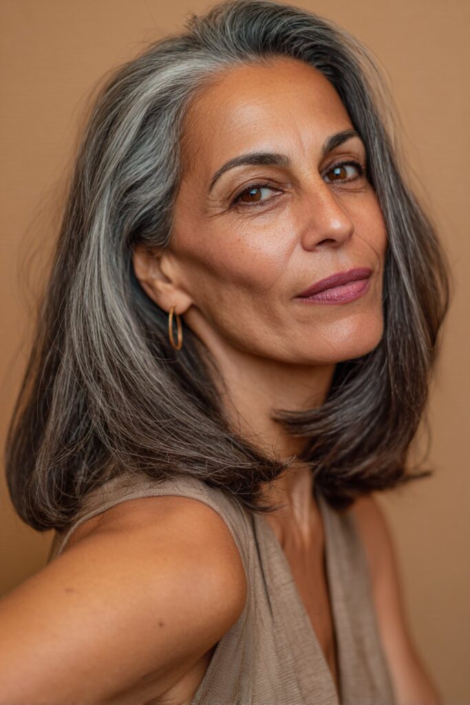 A vibrant elegant woman over 40 with a mature appearance, showcasing a Medium Length Hairstyle For Over 40 with an angled lob, featuring sharp angled long bob longer in front shorter in back, captured in a close-up shot focusing on the hair, photographed against a seamless warm taupe backdrop with a silky finish. She has subtle makeup with visible aging grace, a chic outfit, realistic hair texture with natural highlights, warm skin tone, and a confident smile, captured with soft natural lighting and an Instagram-style beauty vibe.