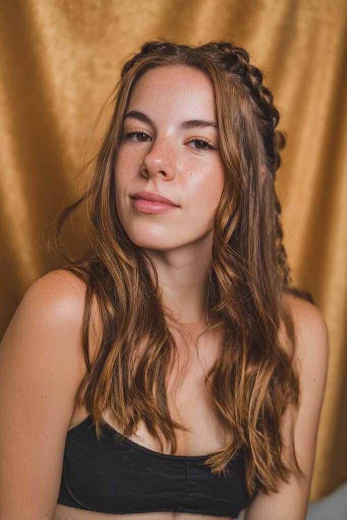 A stunning young woman in her 20s with natural skin tone, showcasing an Everyday Hairstyle For Medium Length Hair with a braided crown accent, featuring medium-length hair with a small braided crown detail, captured in a close-up shot focusing on the hair, photographed against a seamless warm gold backdrop with a glossy sheen. She has subtle makeup, a trendy outfit, realistic hair texture, warm skin tone, and a friendly expression, captured with soft natural lighting and an Instagram-style beauty vibe.