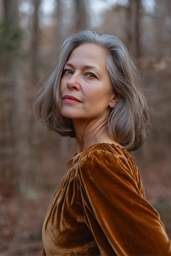A gorgeous older woman over 50 with fine hair styled in a caramel-balayage medium shag, dressed in a full-sleeve velvet blouse, posing in natural light with a forest-themed background.