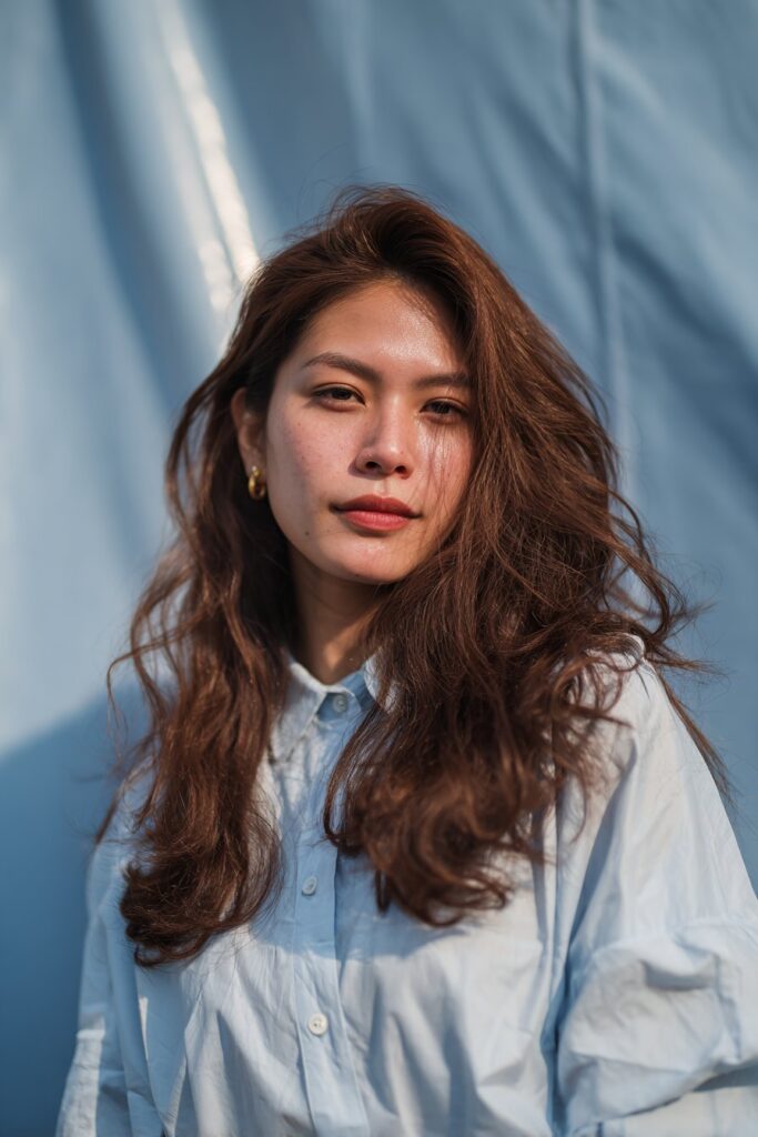 A stunning young woman in her 20s with natural skin tone, showcasing a Winter Hairstyle For Medium Length Hair featuring classic layers with volume, with medium-length hair with classic layered cut and voluminous blowout, captured in a close-up shot focusing on the hair, photographed against a seamless soft navy backdrop with a glossy sheen. She has subtle makeup, a chic winter outfit, realistic hair texture, warm skin tone, and a confident expression, captured with soft natural lighting and an Instagram-style beauty vibe.