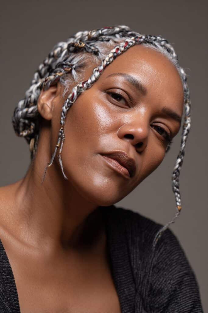 A close-up photo of an elegant Black American woman over 50 with cornrows with white and red highlights hairstyle, featuring realistic hair texture under soft natural lighting, subtle makeup, dark skin tone, confident expression, fully clothed in a chic outfit, in an Instagram-style beauty shot.