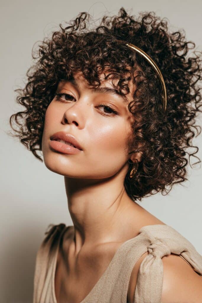 A close-up photo of a young elegant American woman with a short curly bob enhanced by a slim metallic headband, featuring realistic curly texture under soft natural lighting, subtle makeup, warm skin tone, confident expression, fully clothed in a chic outfit, in an Instagram-style beauty shot – headband hairstyles curly hair series.