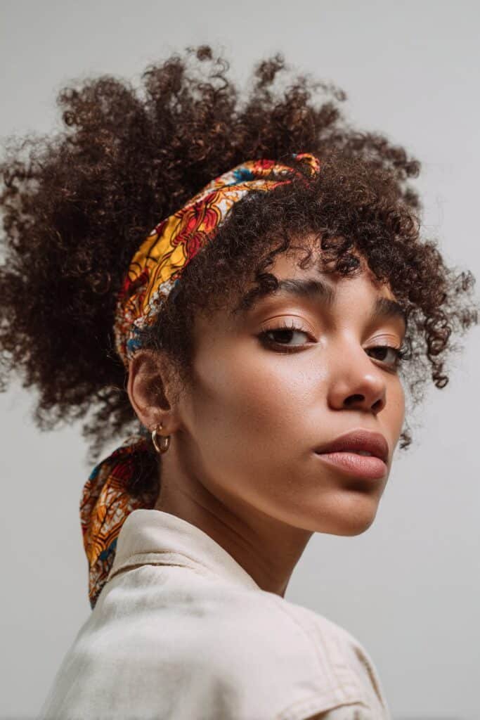 A close-up photo of a young elegant Black American woman with a full curly afro styled with an African print headband, featuring realistic curly afro texture under soft natural lighting, subtle makeup, warm skin tone, confident expression, fully clothed in a chic outfit, in an Instagram-style beauty shot – headband hairstyles black women series.
