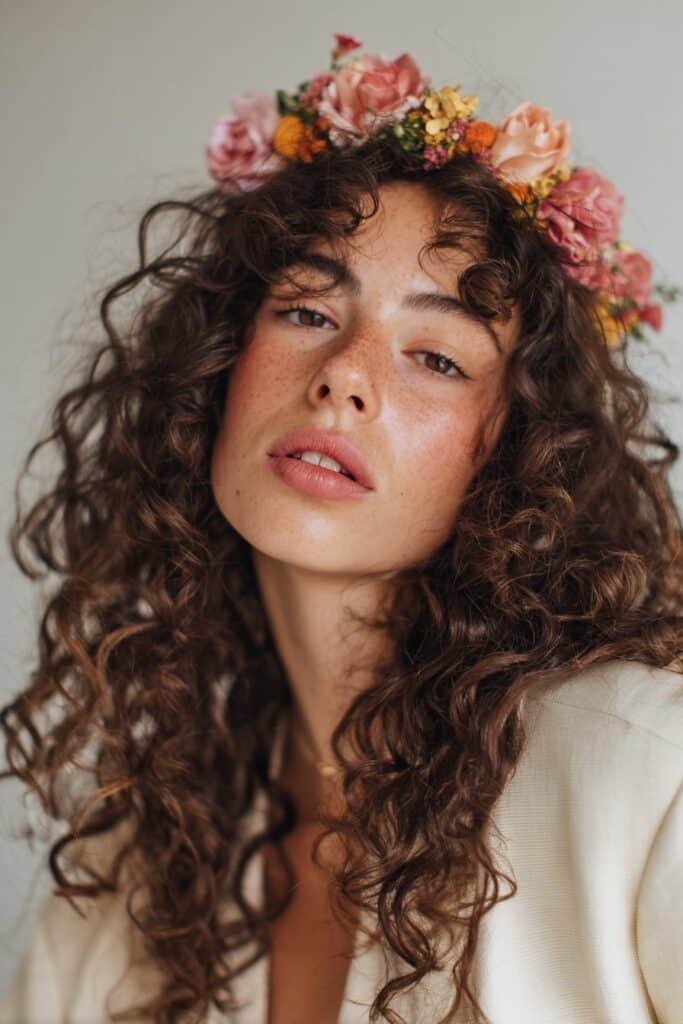 A close-up photo of a young elegant American woman with flowing curls featuring a vibrant floral headband, featuring realistic curly texture under soft natural lighting, subtle makeup, warm skin tone, confident expression, fully clothed in a chic outfit, in an Instagram-style beauty shot – headband hairstyles curly hair series.