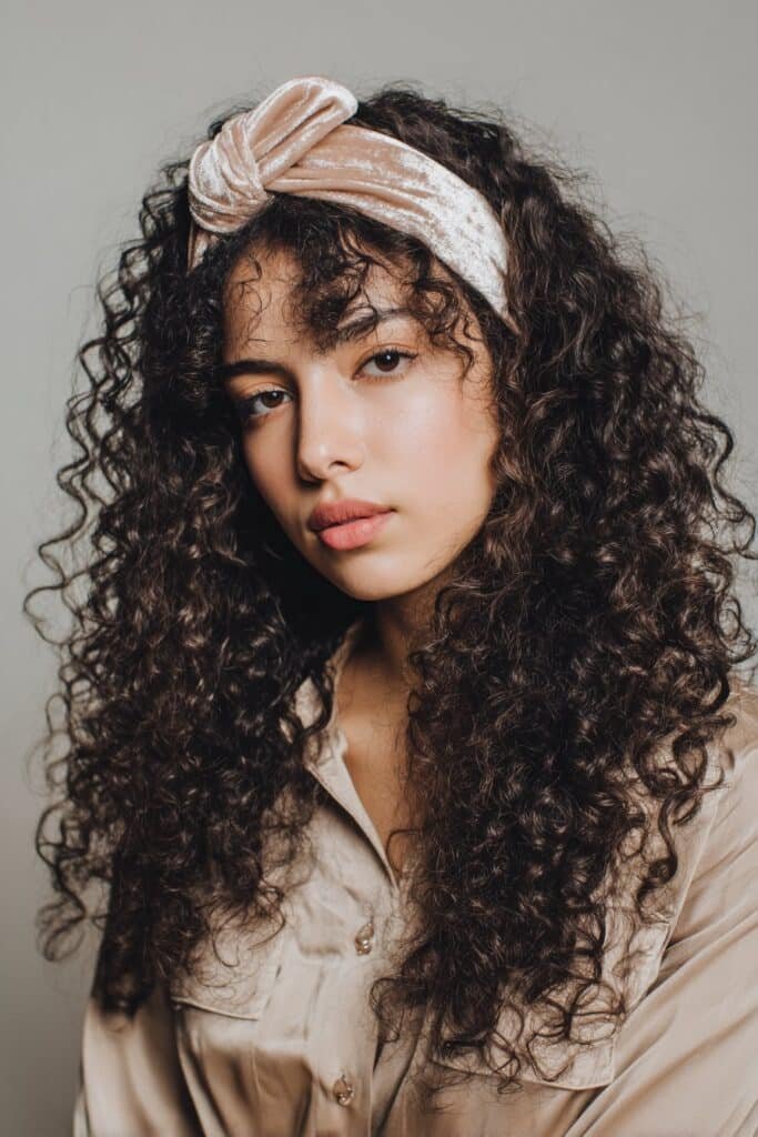 A close-up photo of a young elegant American woman with luxurious curls accented by a plush velvet headband, featuring realistic curly texture under soft natural lighting, subtle makeup, warm skin tone, confident expression, fully clothed in a chic outfit, in an Instagram-style beauty shot – headband hairstyles curly hair series.