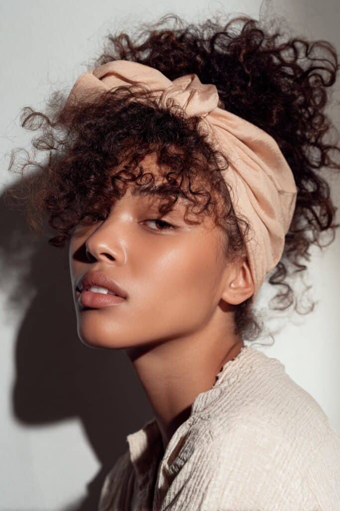 A close-up photo of a young elegant American woman with voluminous curls pushed back by a thick wide headband, featuring realistic curly texture under soft natural lighting, subtle makeup, warm skin tone, confident expression, fully clothed in a chic outfit, in an Instagram-style beauty shot – headband hairstyles curly hair series.