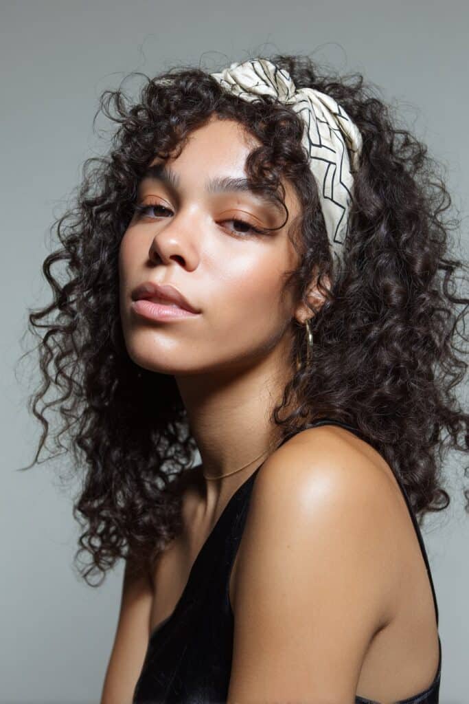 A close-up photo of a young elegant American woman with playful curls secured by a zig-zag headband, featuring realistic curly texture under soft natural lighting, subtle makeup, warm skin tone, confident expression, fully clothed in a chic outfit, in an Instagram-style beauty shot – headband hairstyles curly hair series.
