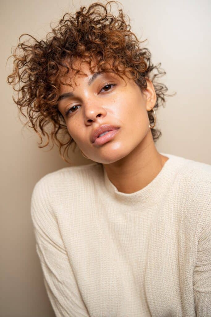 A curly mixie haircut combining a pixie and a mullet, featuring tight natural curls with very short micro bangs. The textured curls frame the forehead while the soft shaggy layers add movement and shape around the face.