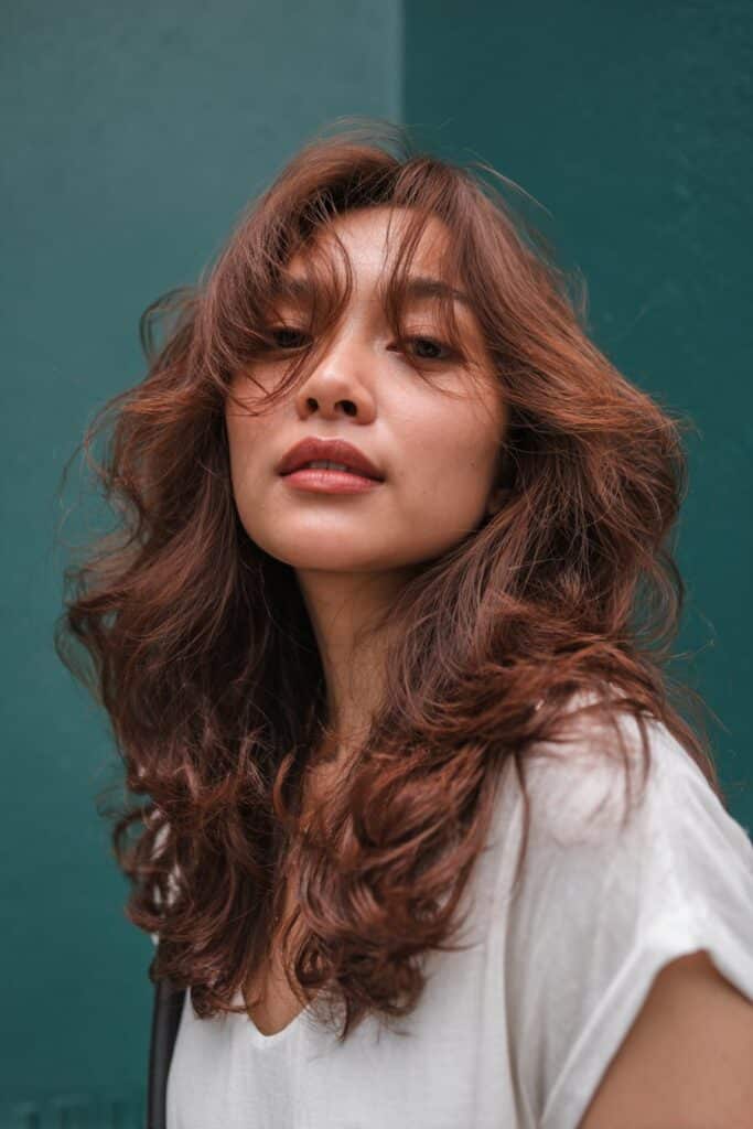 A stunning young woman in her 20s with natural skin tone, showcasing a Brown Hair With Caramel Highlights hairstyle featuring dimensional chocolate copper, featuring chocolate brown hair with dimensional copper and caramel highlights, captured in a close-up shot focusing on the hair, photographed against a seamless deep emerald backdrop with a satin finish. She has subtle makeup, a chic outfit, realistic hair texture, warm skin tone, and a confident expression, captured with soft natural lighting and an Instagram-style beauty vibe.