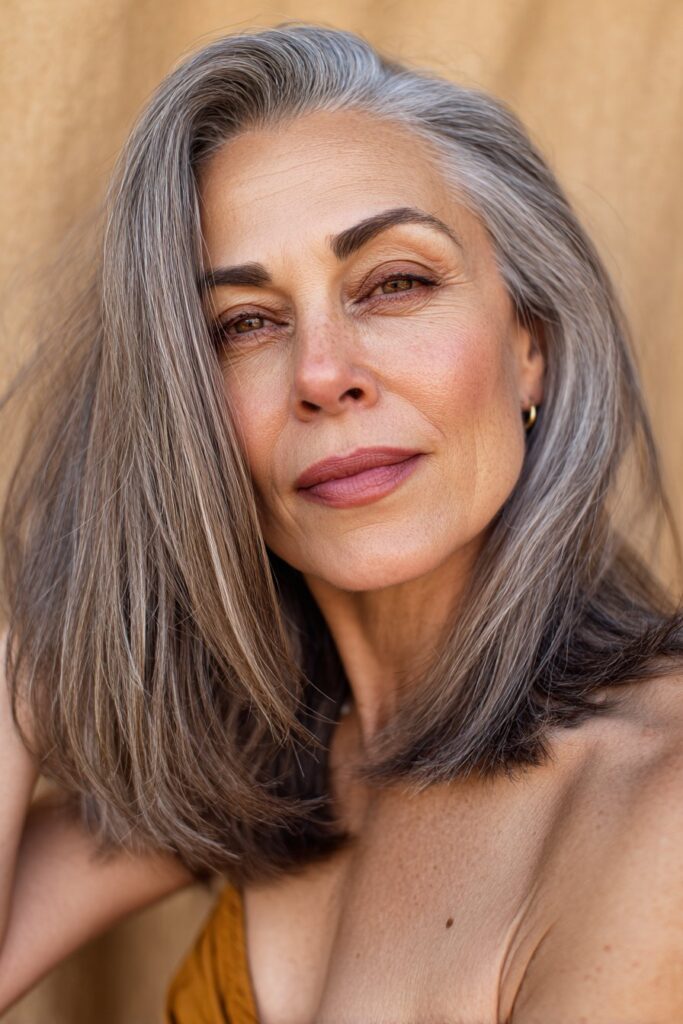 A vibrant American female model over 60 with a mature appearance, showcasing a Medium Length Layered Haircut For Women Over 60 with a feathered shoulder cut, featuring medium-length shoulder cut with feathered layers for movement, captured in a close-up shot focusing on the hair, photographed against a seamless warm beige backdrop with a silky finish. She has subtle makeup with visible aging signs, an elegant outfit, realistic hair texture with silver strands, warm skin tone, and a friendly expression, captured with soft natural lighting and an Instagram-style beauty vibe.