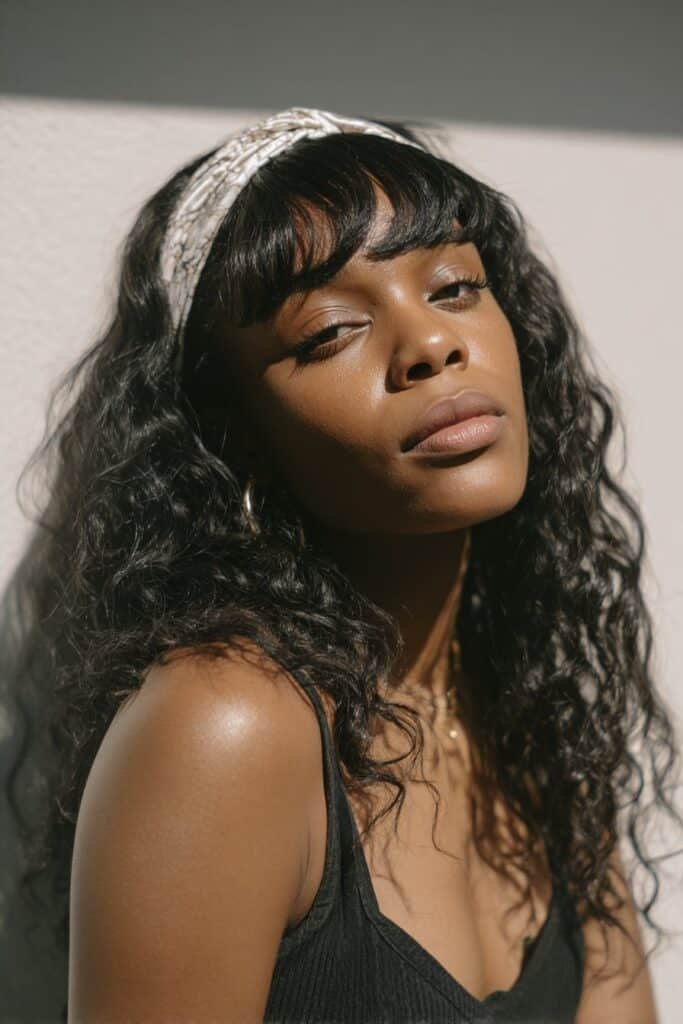 A close-up photo of a young elegant Black American woman with blunt front bangs and a wide headband, featuring realistic hair texture under soft natural lighting, subtle makeup, warm skin tone, confident expression, fully clothed in a chic outfit, in an Instagram-style beauty shot – headband hairstyles black women series.