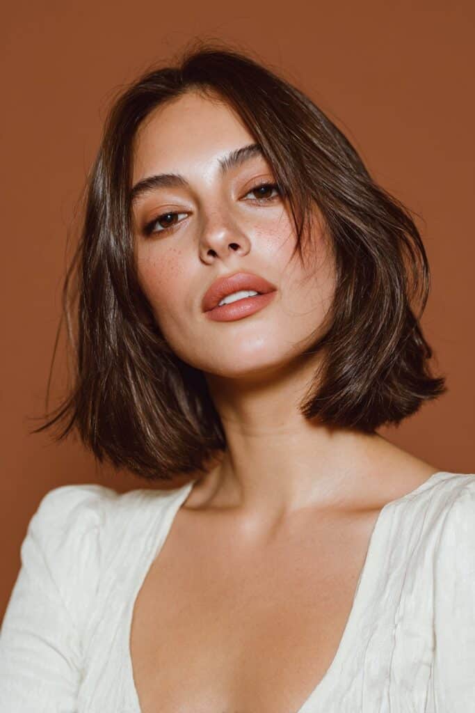 A stunning young woman in her 20s with natural skin tone, showcasing a Winter Hairstyle For Medium Length Hair featuring a glossy brunette bob, with medium-length glossy brunette bob with sleek shine and tucked ends, captured in a close-up shot focusing on the hair, photographed against a seamless warm bronze backdrop with a satin finish. She has subtle makeup, a chic winter outfit, realistic hair texture, warm skin tone, and a confident expression, captured with soft natural lighting and an Instagram-style beauty vibe.