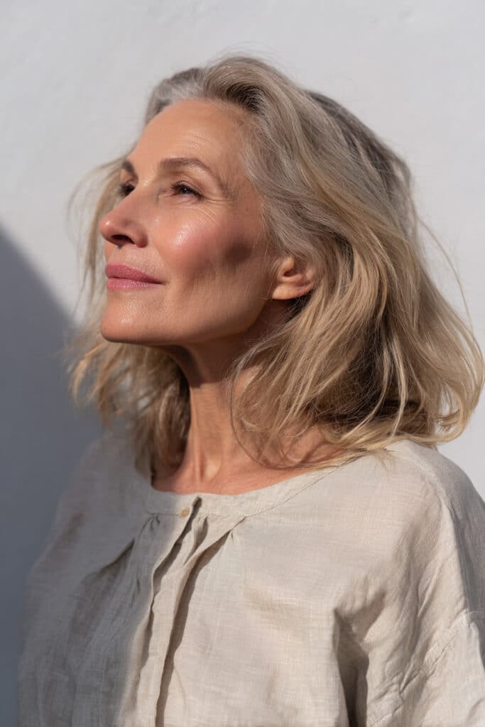 A radiant older woman over 50 with fine golden-blonde wavy shag hair, wearing a full-sleeve linen blouse, photographed in soft natural lighting against a seamless white backdrop.