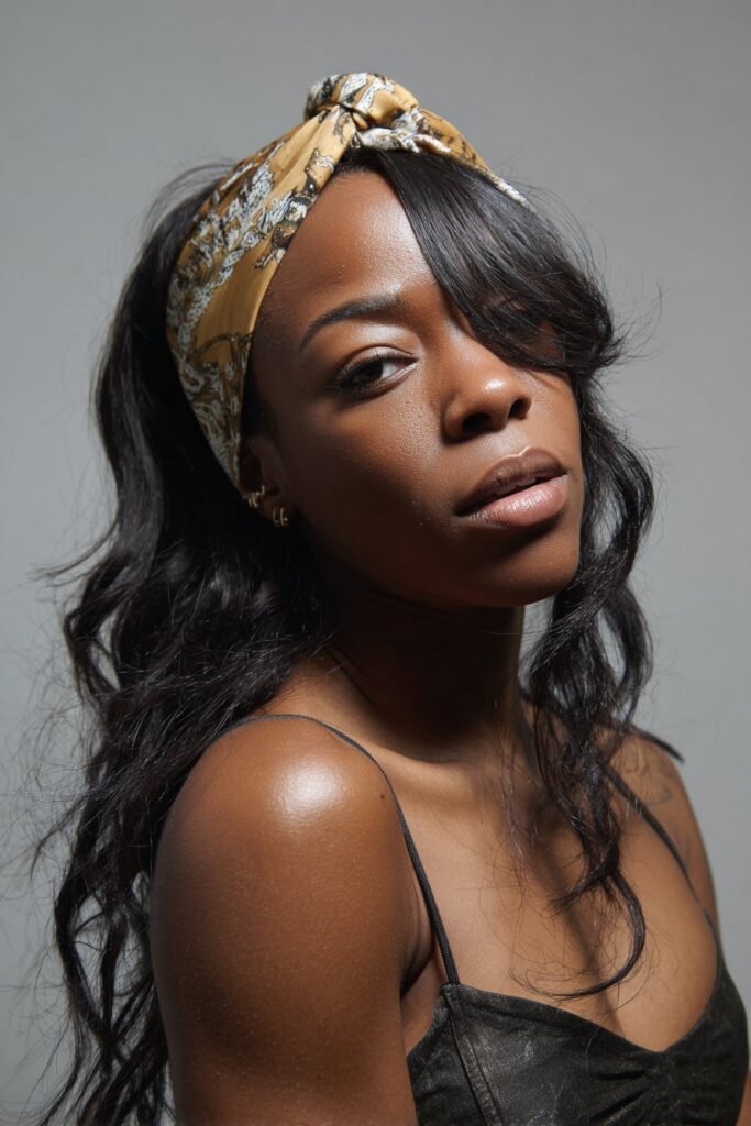 A close-up photo of a young elegant Black American woman with a dramatic swoop bang and slim headband, featuring realistic hair texture under soft natural lighting, subtle makeup, warm skin tone, confident expression, fully clothed in a chic outfit, in an Instagram-style beauty shot – headband hairstyles black women series.