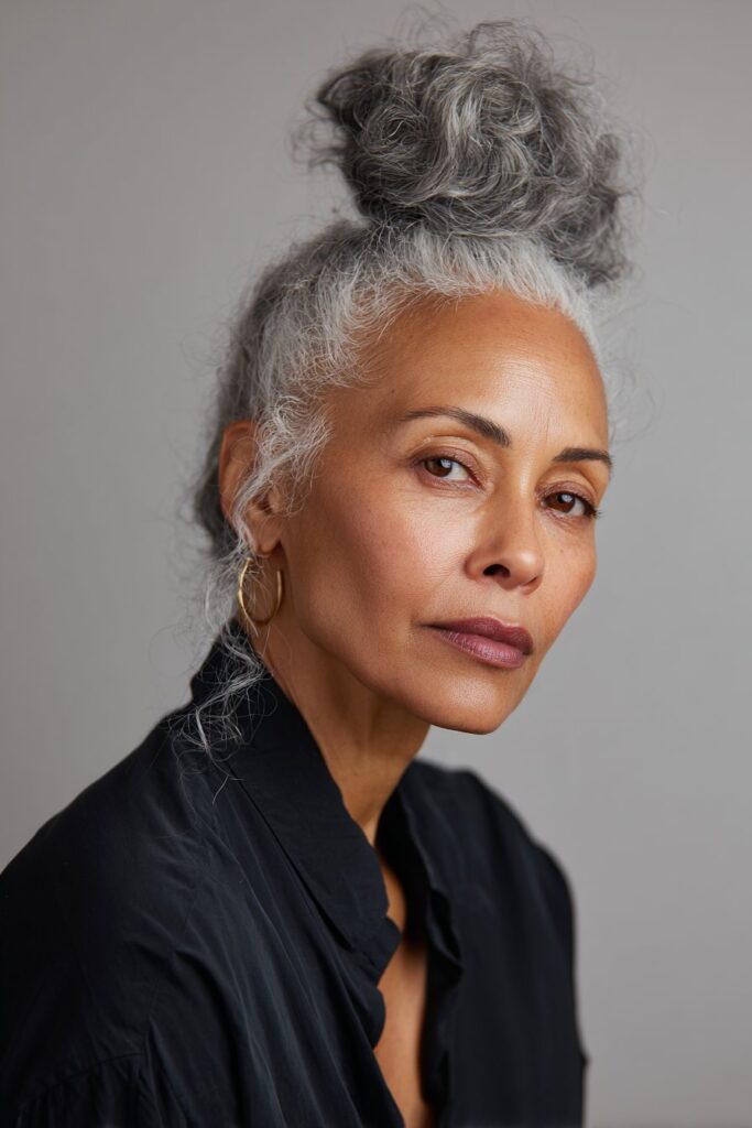 A close-up photo of an elegant Black American woman over 50 with a high bun featuring gray accents hairstyle, realistic hair texture, soft natural lighting, subtle makeup, confident expression, chic outfit, Instagram-style beauty shot.