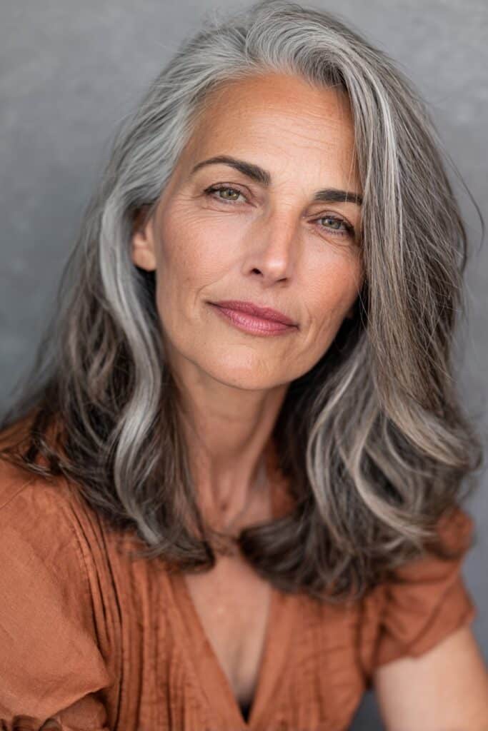 A vibrant American female model over 60 with a mature appearance, showcasing a Medium Length Layered Haircut For Women Over 60 with a layered lob, featuring medium-length lob with soft layers for volume, captured in a close-up shot focusing on the hair, photographed against a seamless soft gray backdrop with a matte finish. She has subtle makeup with visible aging signs, an elegant outfit, realistic hair texture with silver strands, warm skin tone, and a friendly expression, captured with soft natural lighting and an Instagram-style beauty vibe.