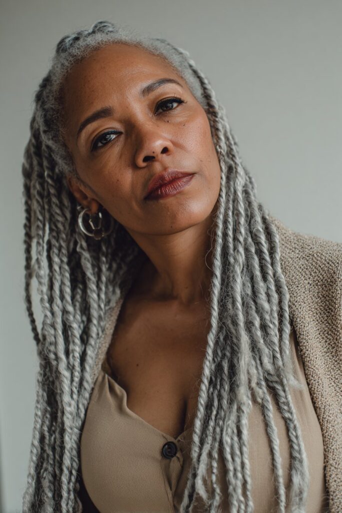 A close-up photo of an elegant Black American woman over 50 with long box braids with gray accents hairstyle, featuring realistic hair texture under soft natural lighting, subtle makeup, dark skin tone, confident expression, fully clothed in a chic outfit, in an Instagram-style beauty shot.