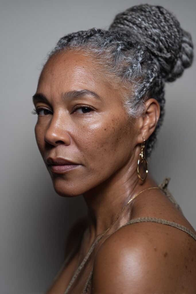 A close-up photo of an elegant Black American woman over 50 with a low braided bun with gray cornrows hairstyle, featuring realistic hair texture under soft natural lighting, subtle makeup, dark skin tone, confident expression, fully clothed in a chic outfit, in an Instagram-style beauty shot.