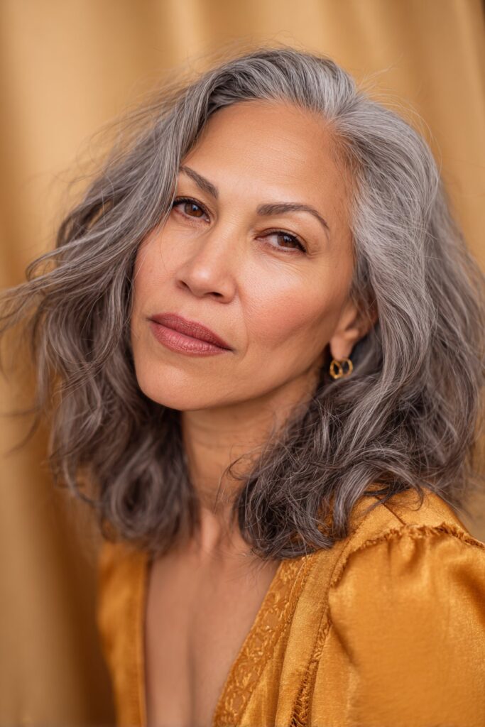 A vibrant American female model over 60 with a mature appearance, showcasing a Medium Length Layered Haircut For Women Over 60 with a low maintenance layered cut, featuring medium-length layered cut with minimal styling for ease, captured in a close-up shot focusing on the hair, photographed against a seamless warm gold backdrop with a glossy sheen. She has subtle makeup with visible aging signs, an elegant outfit, realistic hair texture with silver strands, warm skin tone, and a friendly expression, captured with soft natural lighting and an Instagram-style beauty vibe.