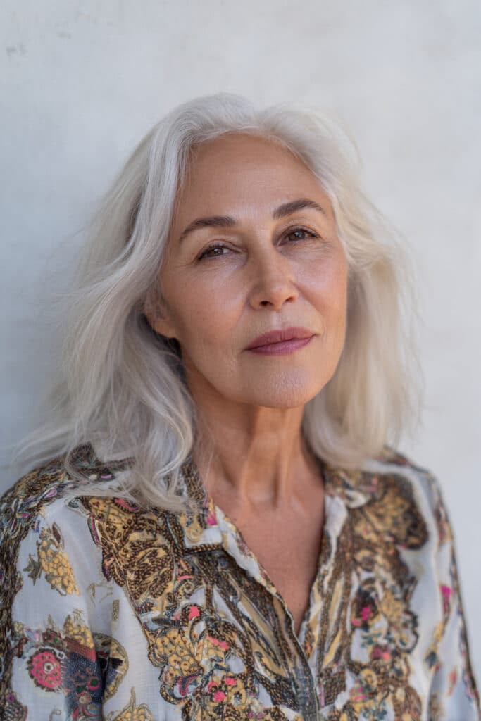 A confident older woman over 50 with fine platinum-blonde shag hair featuring dark roots, dressed in a full-sleeve embroidered shirt, photographed in natural bright light with a seamless white backdrop.
