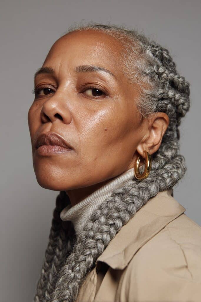 A close-up photo of an elegant Black American woman over 50 with salt-and-pepper box braids hairstyle, featuring realistic hair texture under soft natural lighting, subtle makeup, dark skin tone, confident expression, fully clothed in a chic outfit, in an Instagram-style beauty shot.