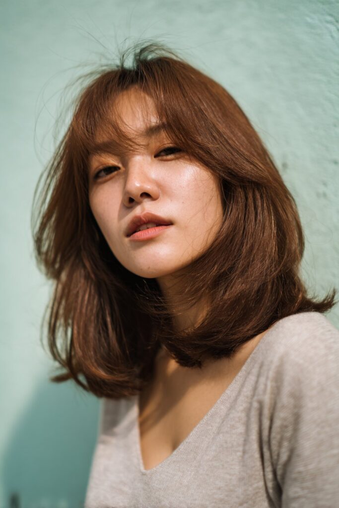 A fashionable American woman in her 20s with a shag haircut and side-swept bangs, tousled and textured for a relaxed look, captured in soft natural light against a light aqua background.