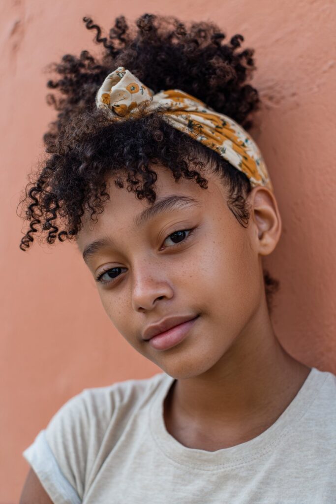 American student with short 4C afro accented by a colorful headband, wearing a button-up shirt, smiling outdoors on the school grounds.