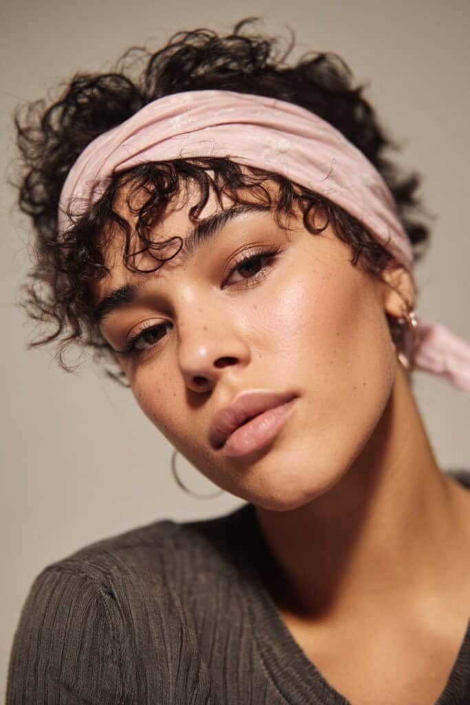 A close-up photo of a young elegant American woman with short curly hair gently held back by a thin headband, featuring realistic curly texture under soft natural lighting, subtle makeup, warm skin tone, confident expression, fully clothed in a chic outfit, in an Instagram-style beauty shot – headband hairstyles curly hair series.