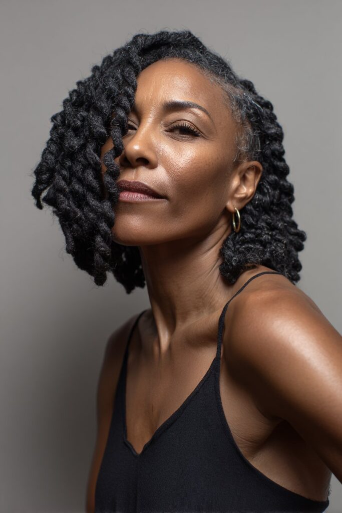A close-up photo of an elegant Black American woman over 50 with a shoulder-length braided bob hairstyle, featuring realistic hair texture under soft natural lighting, subtle makeup, warm skin tone, confident expression, fully clothed in a chic outfit, in an Instagram-style beauty shot.
