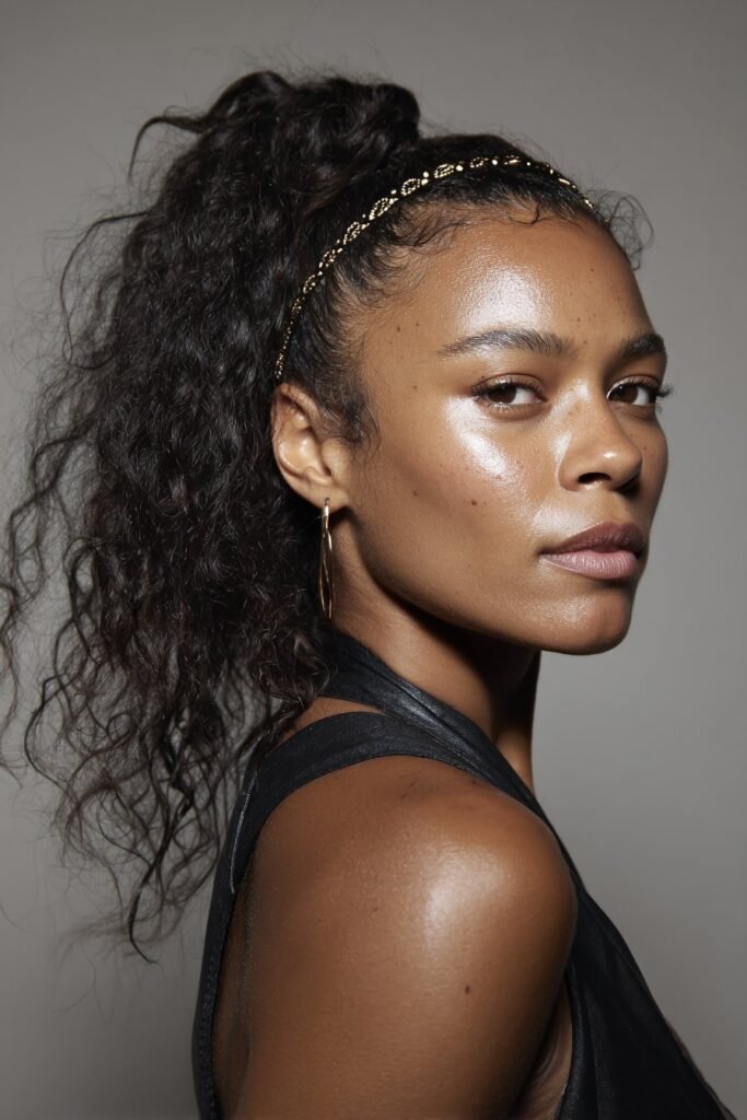 A close-up photo of a young elegant Black American woman with a low side ponytail accented by a narrow headband, featuring realistic hair texture under soft natural lighting, subtle makeup, warm skin tone, confident expression, fully clothed in a chic outfit, in an Instagram-style beauty shot – headband hairstyles black women series.