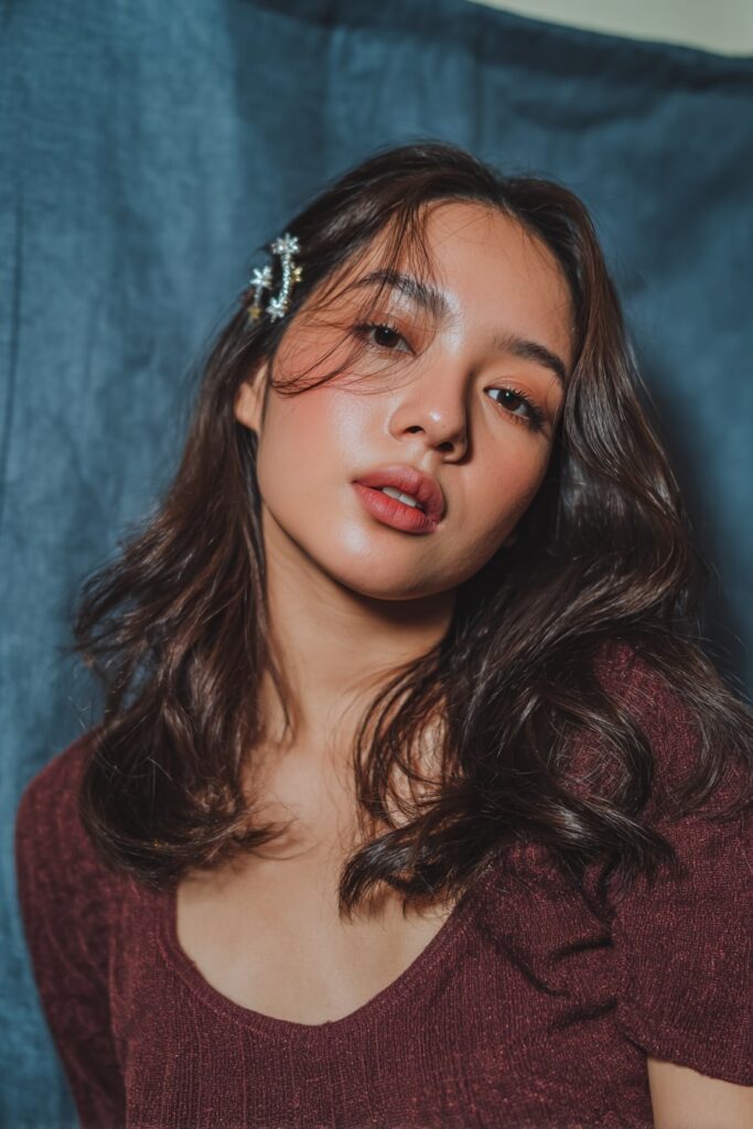 A stunning young woman in her 20s with natural skin tone, showcasing an Everyday Hairstyle For Medium Length Hair with a side sweep with clip, featuring medium-length hair swept to one side with a decorative clip, captured in a close-up shot focusing on the hair, photographed against a seamless soft navy backdrop with a glossy sheen. She has subtle makeup, a trendy outfit, realistic hair texture, warm skin tone, and a friendly expression, captured with soft natural lighting and an Instagram-style beauty vibe.
