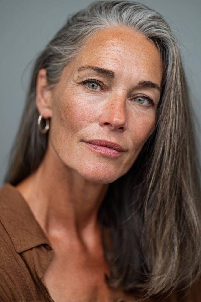 A vibrant elegant woman over 40 with a mature appearance, showcasing a Medium Length Hairstyle For Over 40 with a sleek collarbone cut, featuring sleek straight collarbone-length cut with glass-like shine, captured in a close-up shot focusing on the hair, photographed against a seamless cool slate backdrop with a glossy finish. She has subtle makeup with visible aging grace, a chic outfit, realistic hair texture with natural highlights, warm skin tone, and a confident smile, captured with soft natural lighting and an Instagram-style beauty vibe.