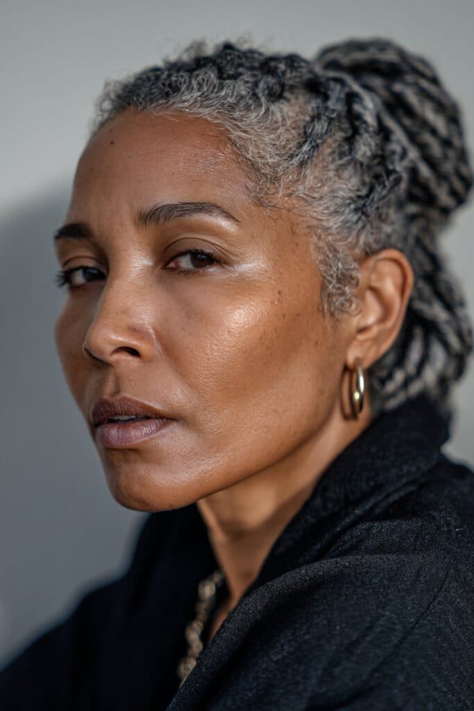 A close-up photo of an elegant Black American woman over 50 with a high bun with gray accents hairstyle, featuring realistic hair texture under soft natural lighting, subtle makeup, dark skin tone, confident expression, fully clothed in a chic outfit, in an Instagram-style beauty shot.