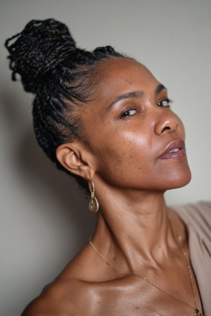 A close-up photo of an elegant Black American woman over 50 with a sleek high bun with micro braids hairstyle, featuring realistic hair texture under soft natural lighting, subtle makeup, dark skin tone, confident expression, fully clothed in a chic outfit, in an Instagram-style beauty shot.