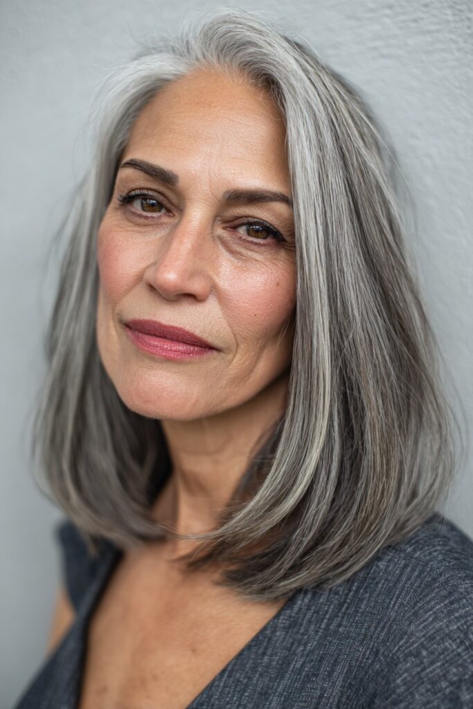 A vibrant American female model over 60 with a mature appearance, showcasing a Medium Length Layered Haircut For Women Over 60 with a sleek layered lob, featuring medium-length sleek layered lob with smooth finish, captured in a close-up shot focusing on the hair, photographed against a seamless light gray backdrop with a smooth surface. She has subtle makeup with visible aging signs, an elegant outfit, realistic hair texture with silver strands, warm skin tone, and a friendly expression, captured with soft natural lighting and an Instagram-style beauty vibe.