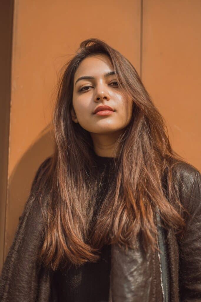 A stunning young woman in her 20s with natural skin tone, showcasing a Winter Hairstyle For Medium Length Hair featuring a sleek winter lob, with medium-length sleek lob with straight polished finish, captured in a close-up shot focusing on the hair, photographed against a seamless warm cinnamon backdrop with a silky finish. She has subtle makeup, a chic winter outfit, realistic hair texture, warm skin tone, and a confident expression, captured with soft natural lighting and an Instagram-style beauty vibe.